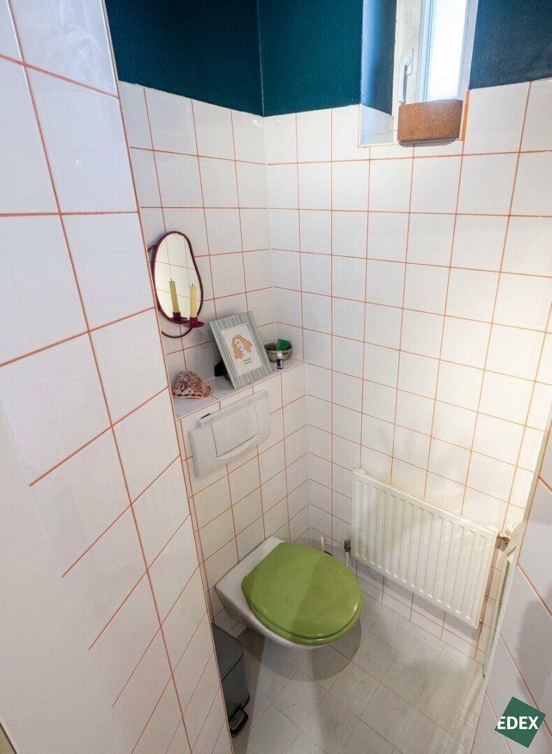 Separate Toilette mit weißen Fliesen, grünem Toilettensitz und einem kleinen Fenster für Belüftung.