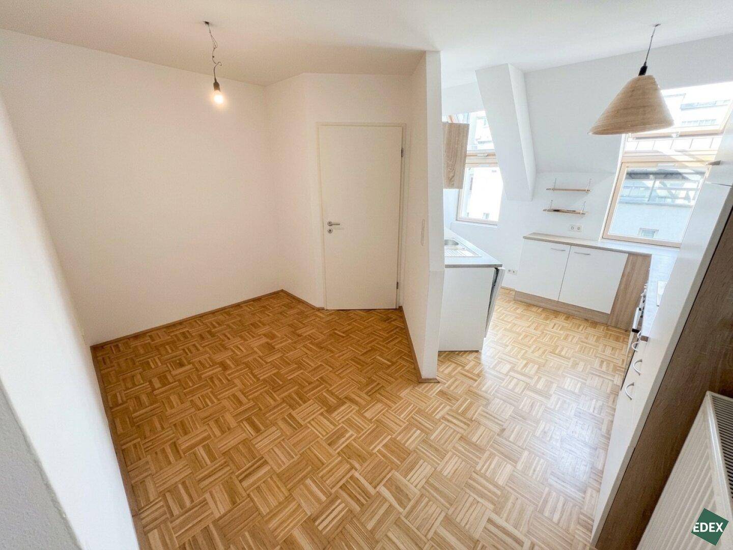Offener Bereich mit Parkettboden und Blick auf eine kleine Küche im Dachgeschoss mit Dachfenstern.