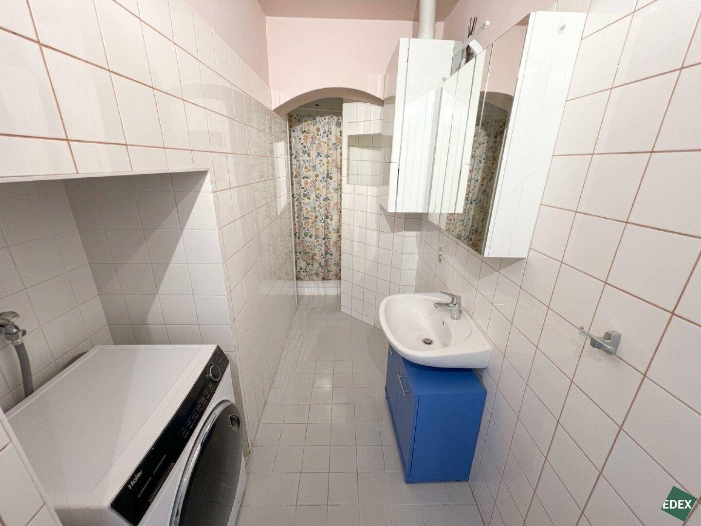 Gefliestes Badezimmer mit Waschbecken, Waschmaschine und Duschbereich mit Vorhang.