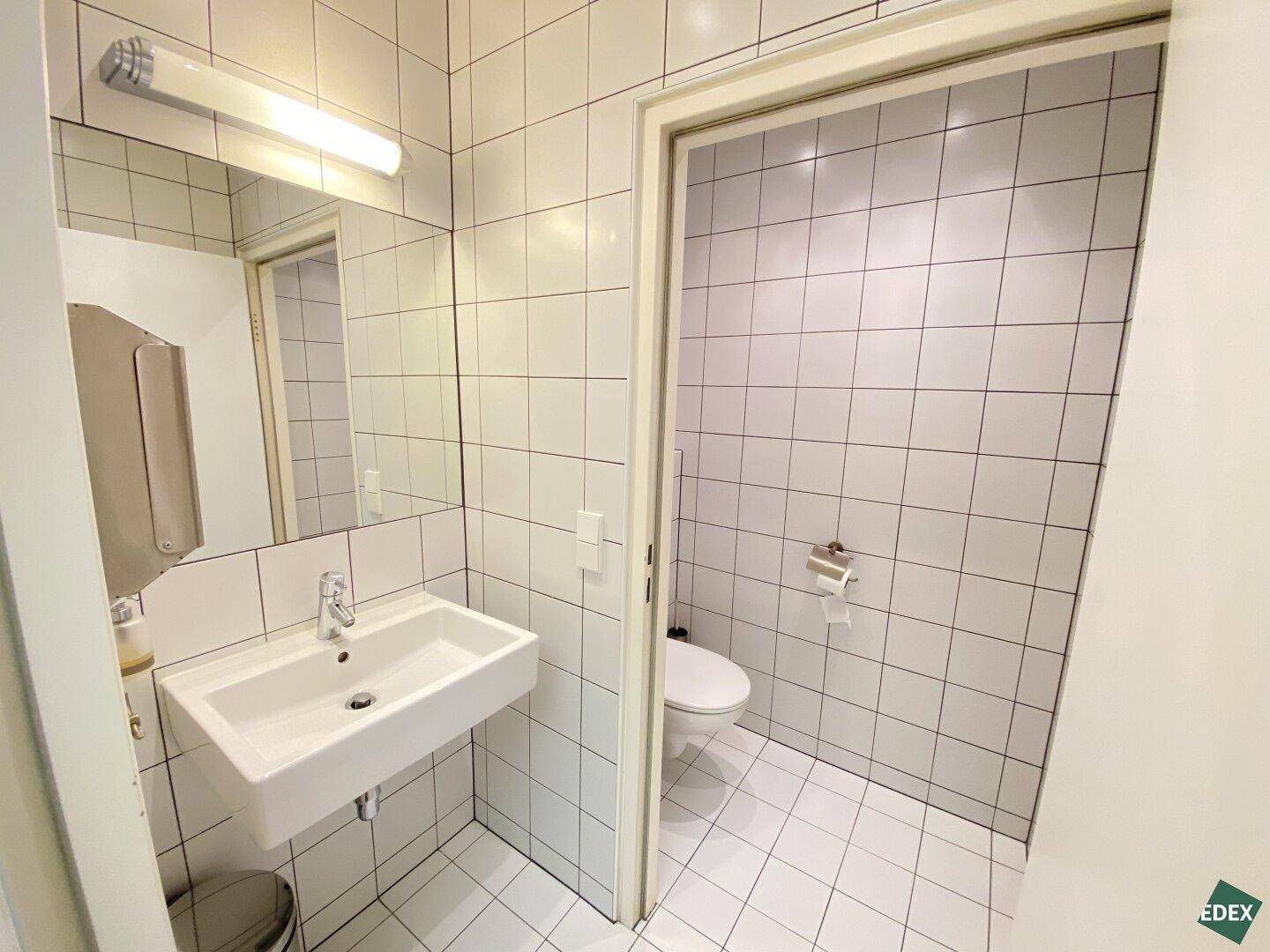 Badezimmer mit weißen Fliesen an Wänden und Boden, einem Waschbecken und einer separaten Toilettennische.
