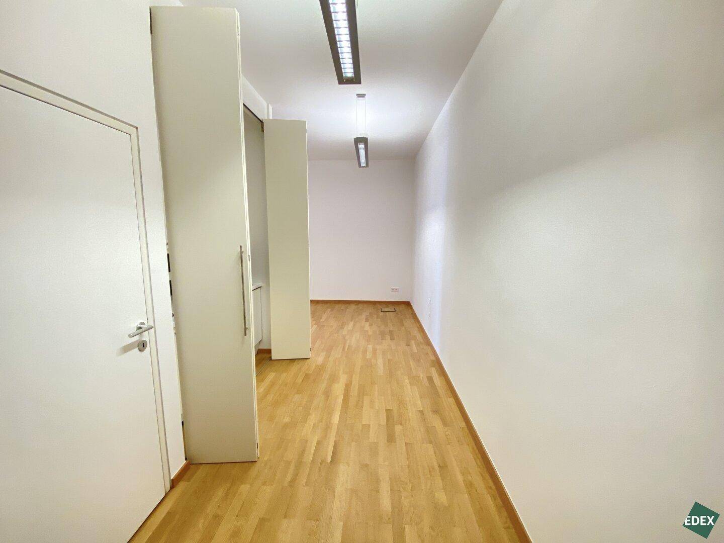 Langer, leerer Büroraum mit hellem Holzparkett, weißen Wänden und moderner Deckenbeleuchtung.