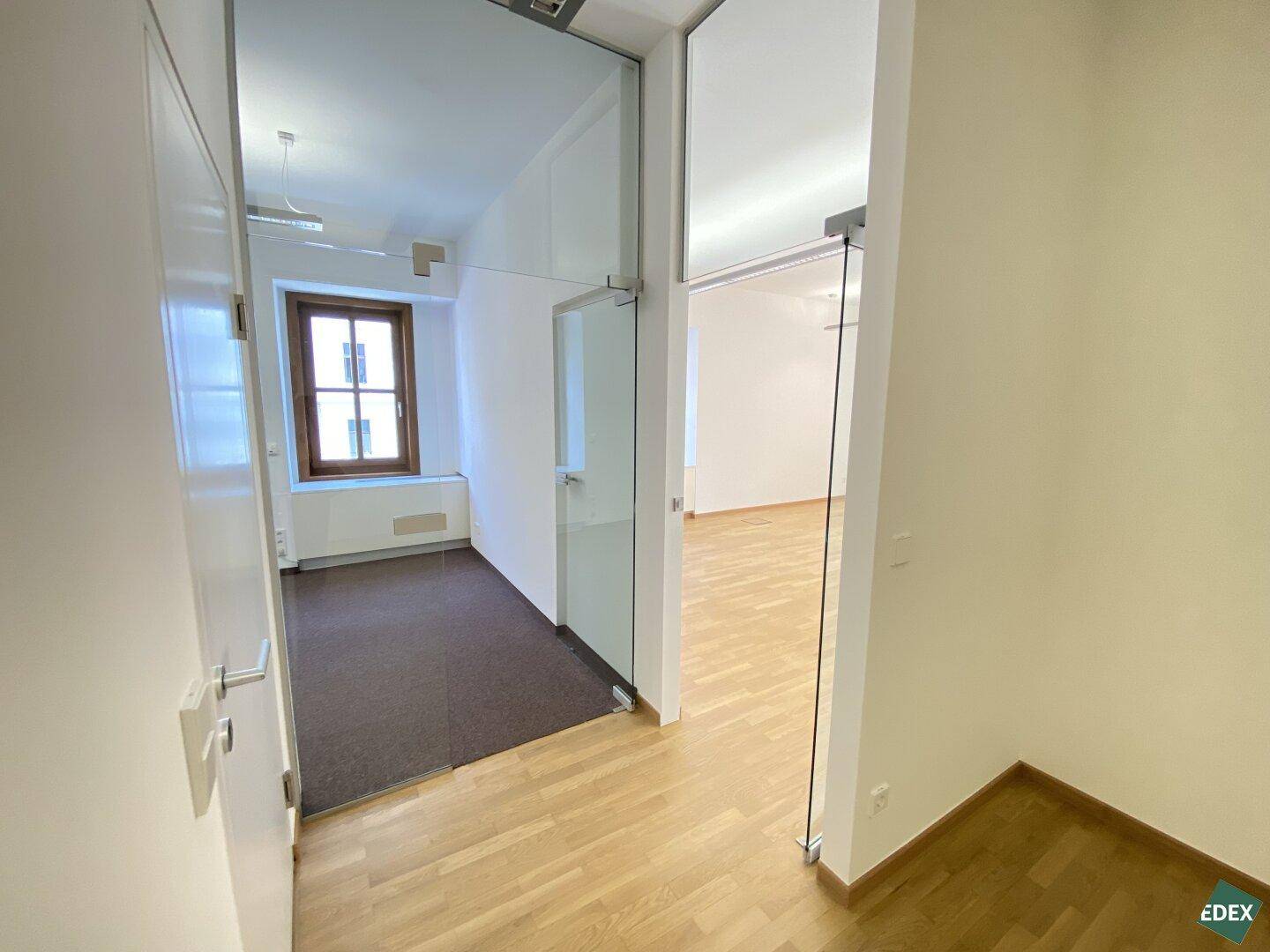Blick durch eine Glastür in einen weiteren Büroraum mit Fenster und Teppichboden, im Vordergrund Holzparkett.