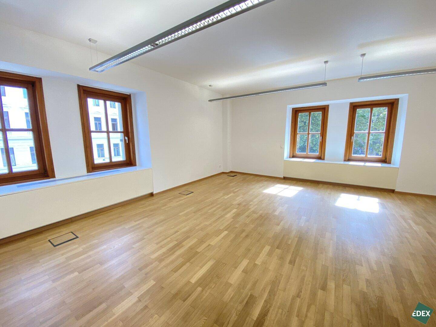Großer, heller Büroraum mit Holzparkett, weißen Wänden und drei großen Fenstern, die viel Licht hereinlassen.