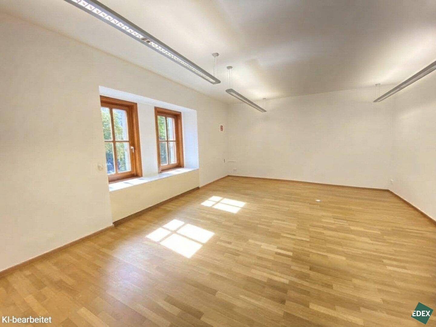 Heller, geräumiger Büroraum mit Holzparkett, weißen Wänden und zwei großen Fenstern, die Sonnenlicht hereinlassen.