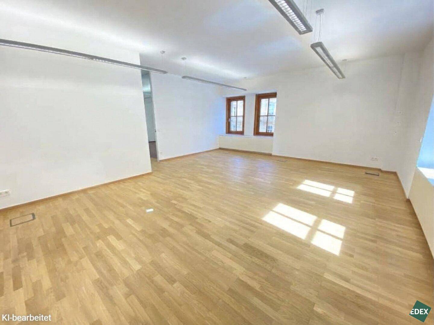 Geräumiger Büroraum mit hellem Holzparkett, weißen Wänden und großen Fenstern, die den Raum mit Licht füllen.