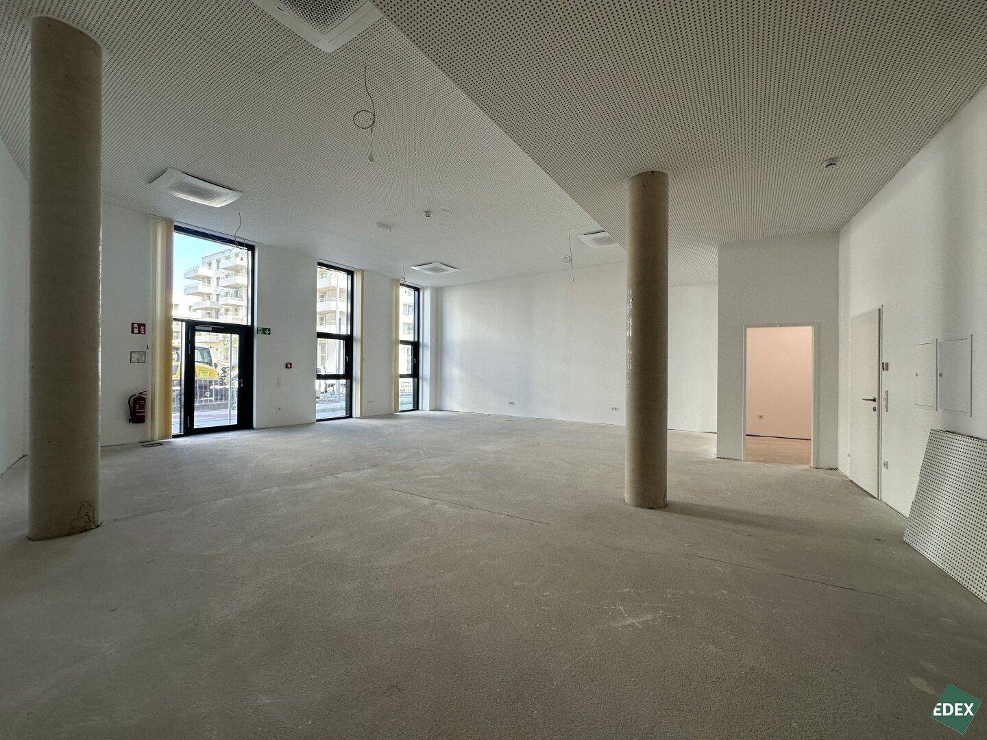 Geräumiger, leerer Gewerberaum mit Betonboden, weißen Wänden, zwei Säulen und großen Fenstern.