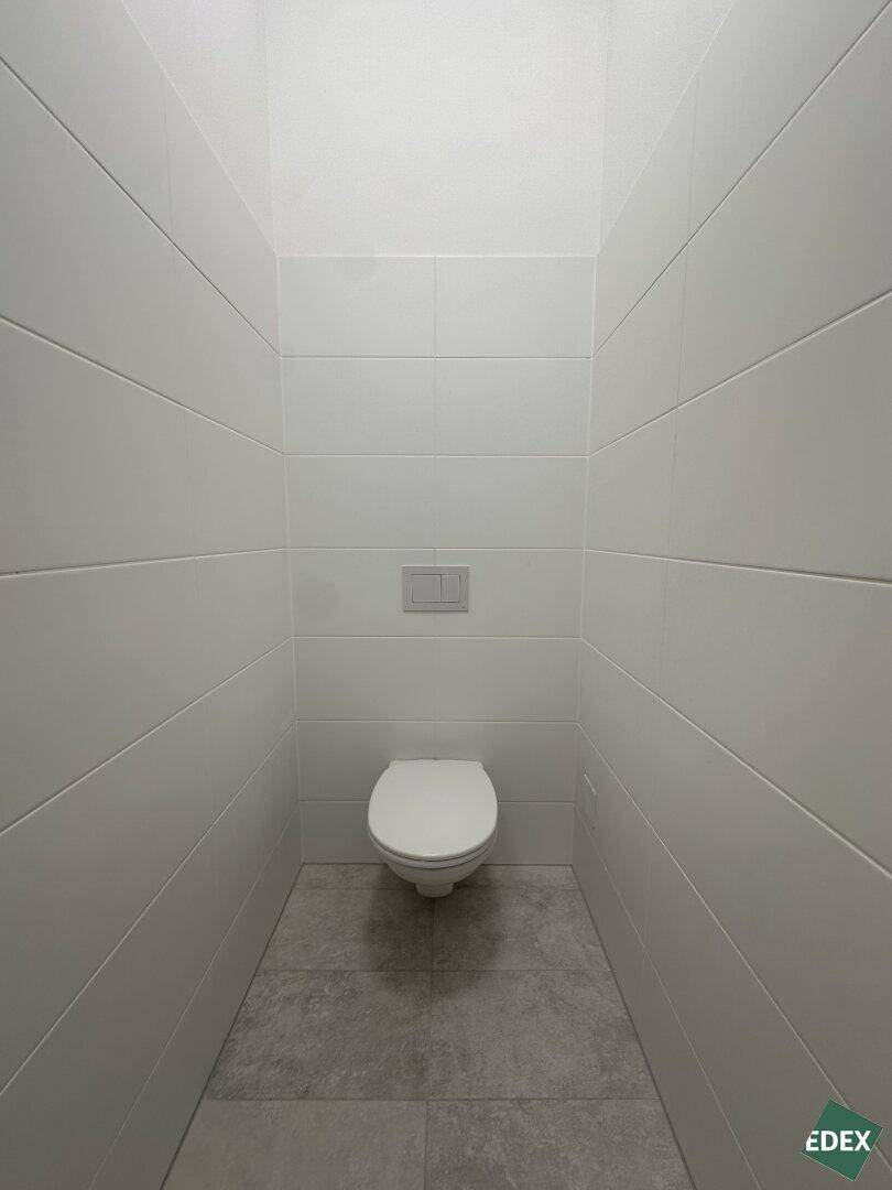 Schmale Toilettenkabine mit wandhängender Toilette, vollständig mit weißen Fliesen verkleidet.