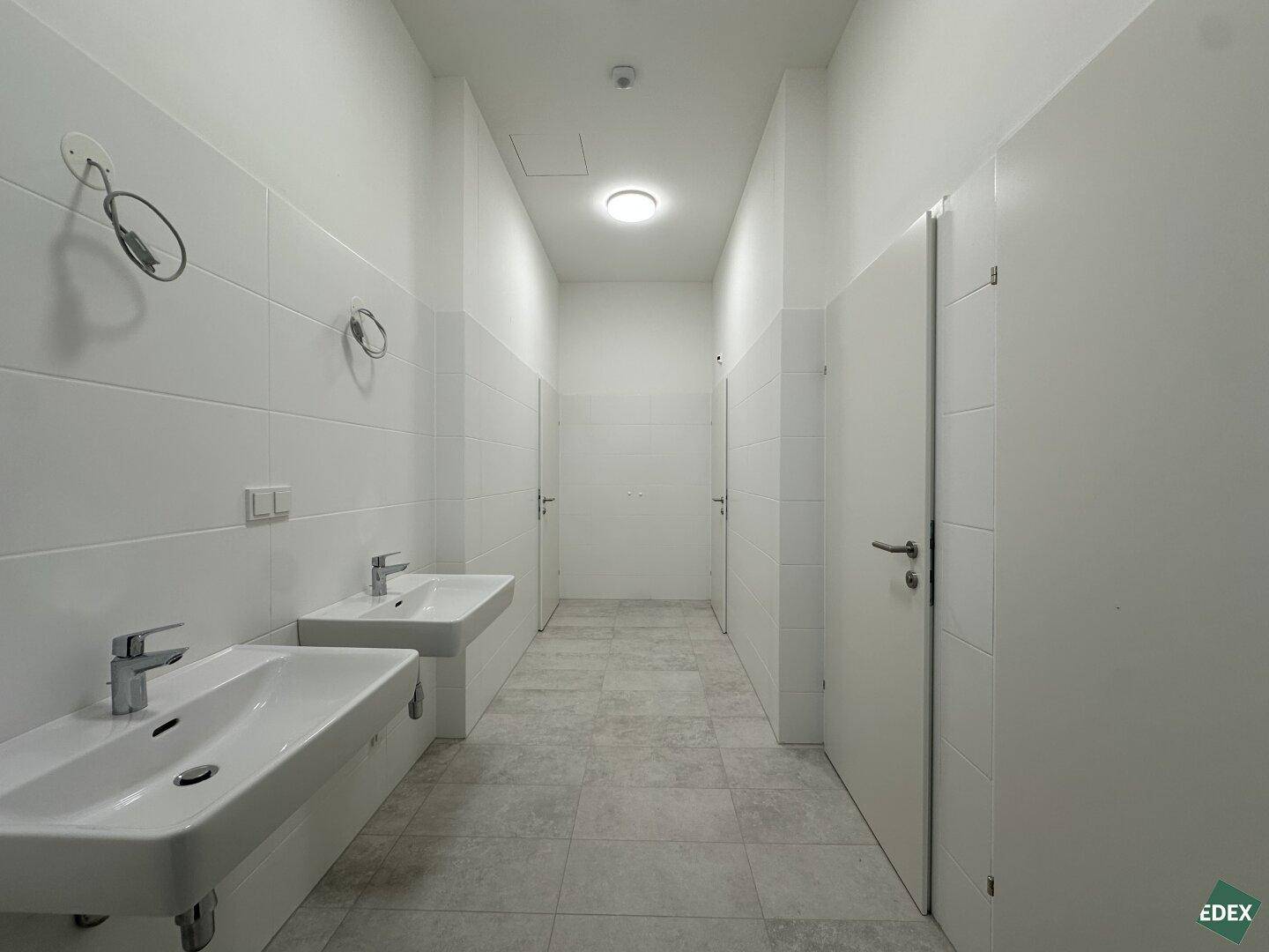 Moderner, gefliester Waschraum mit zwei Waschbecken, weißen Wänden und hellgrauem Fliesenboden.