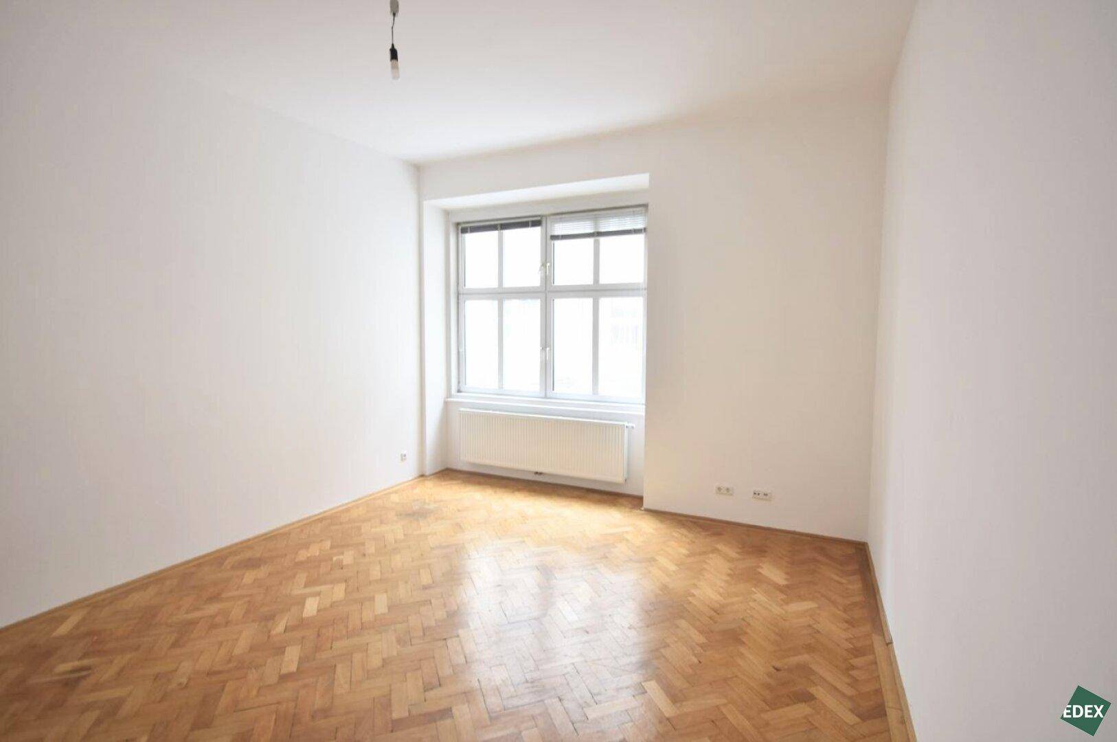 Freundliches Zimmer mit Parkettboden und großem Fenster, das viel Tageslicht hereinlässt.