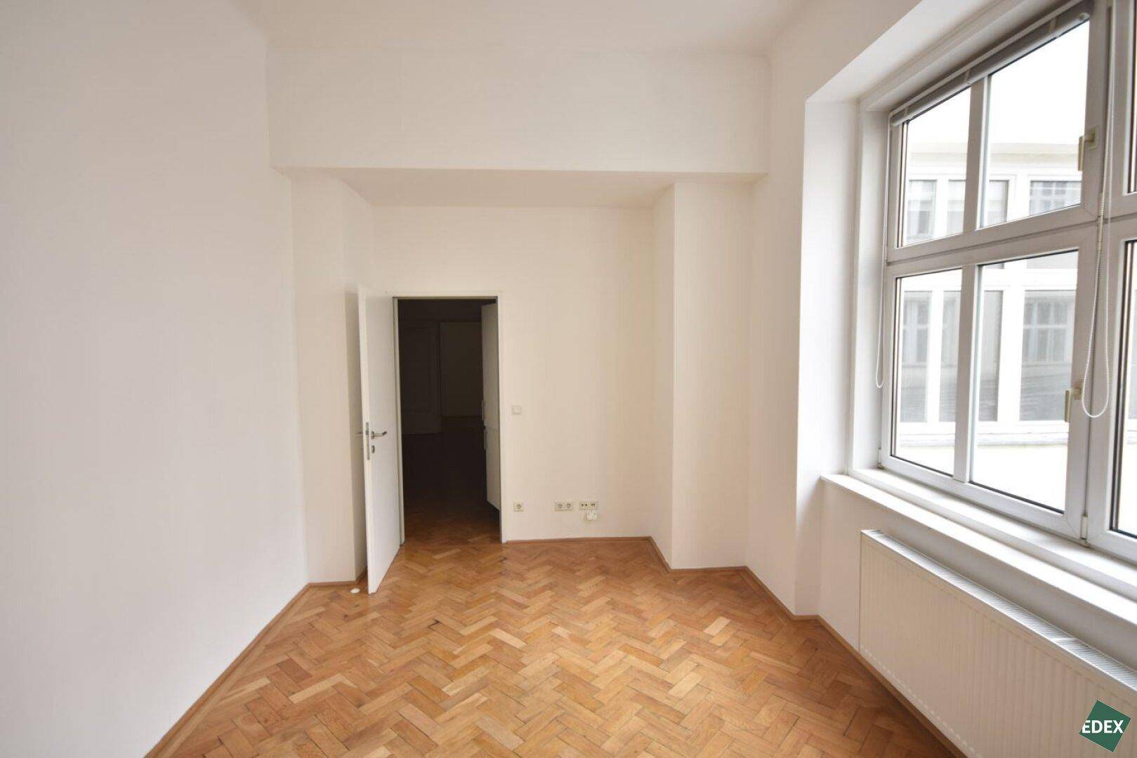 Geräumiges Zimmer mit klassischem Fischgrätparkett, hellen Wänden und Zugang zu weiteren Räumen.