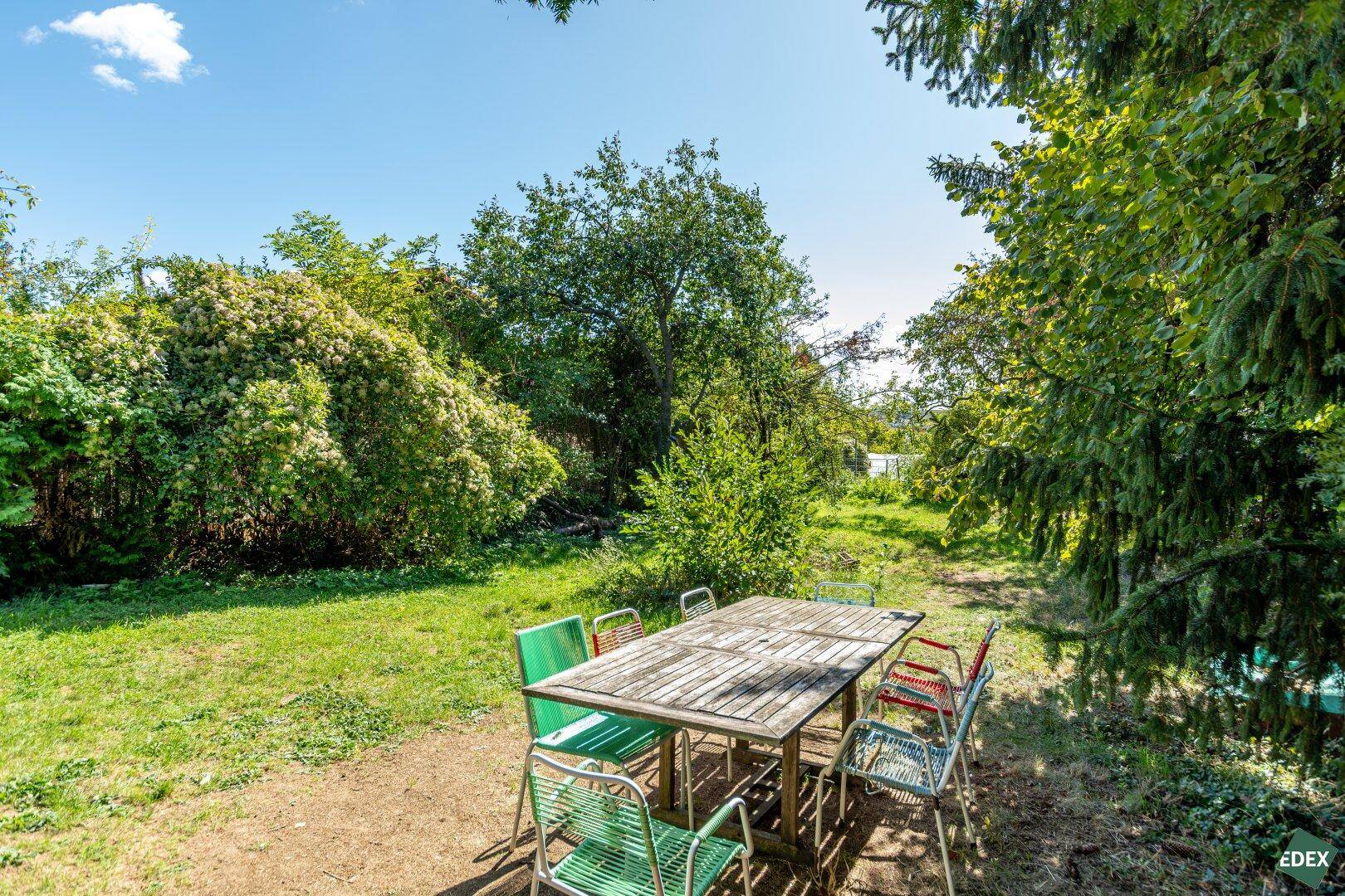 Sonnige Sitzecke im Garten mit rustikalem Holztisch und bunten Stühlen, umgeben von viel Grün.