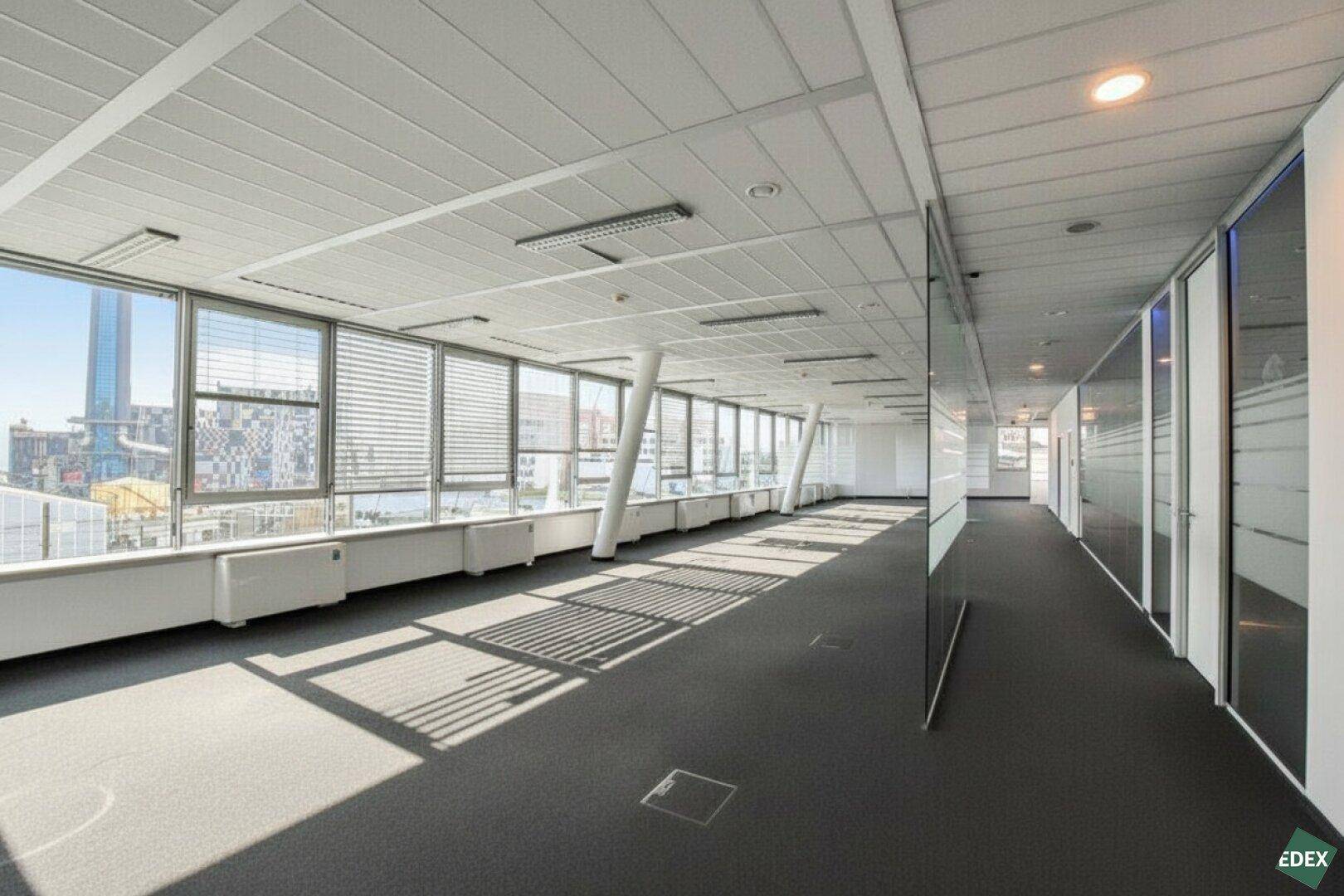 Langer Bürogang mit Glaswänden und großen Fenstern, die einen weiten Stadtblick bieten.