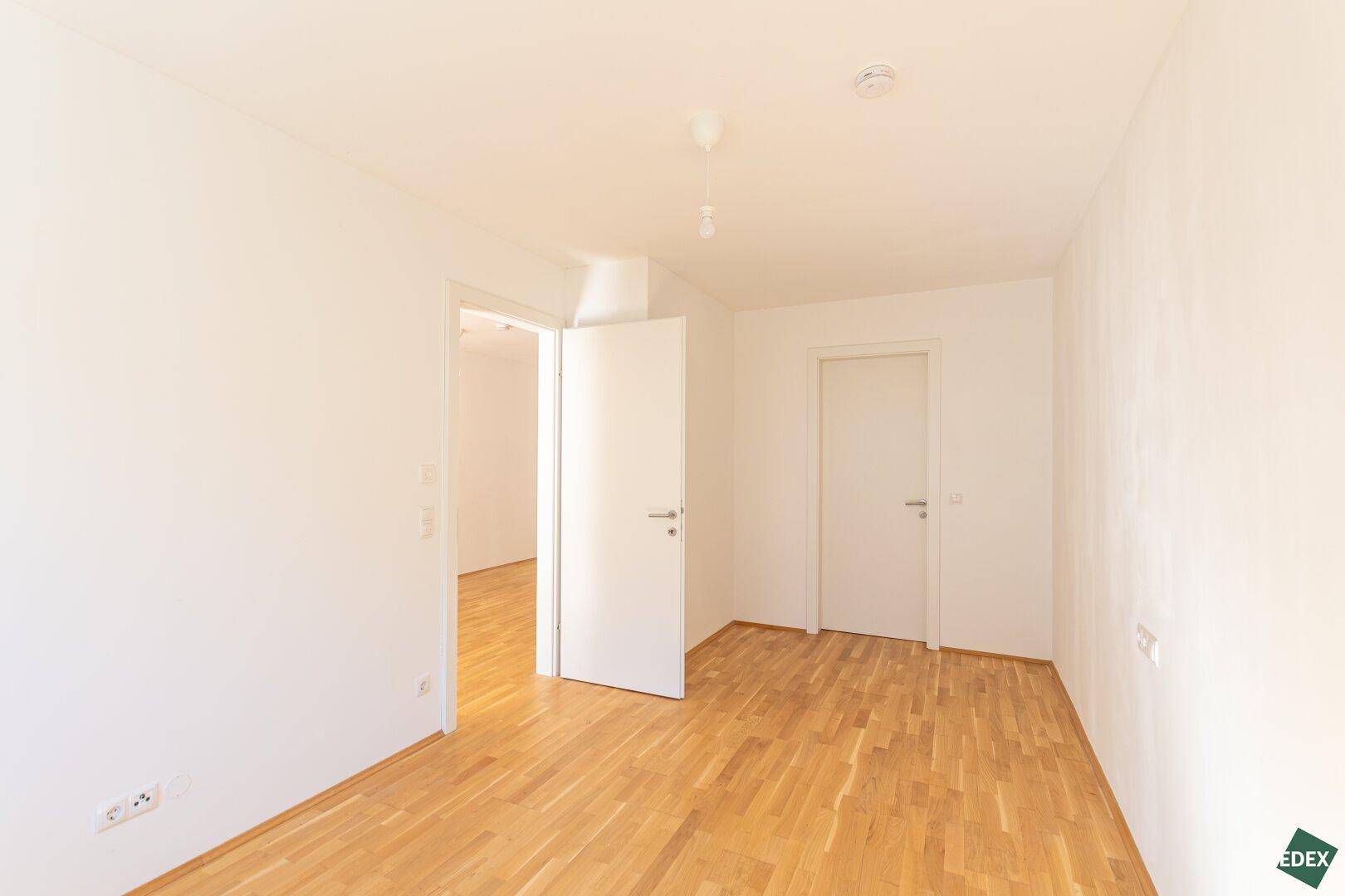 Leeres Zimmer mit Holzboden und weißen Wänden, das durch zwei Türen zugänglich ist.