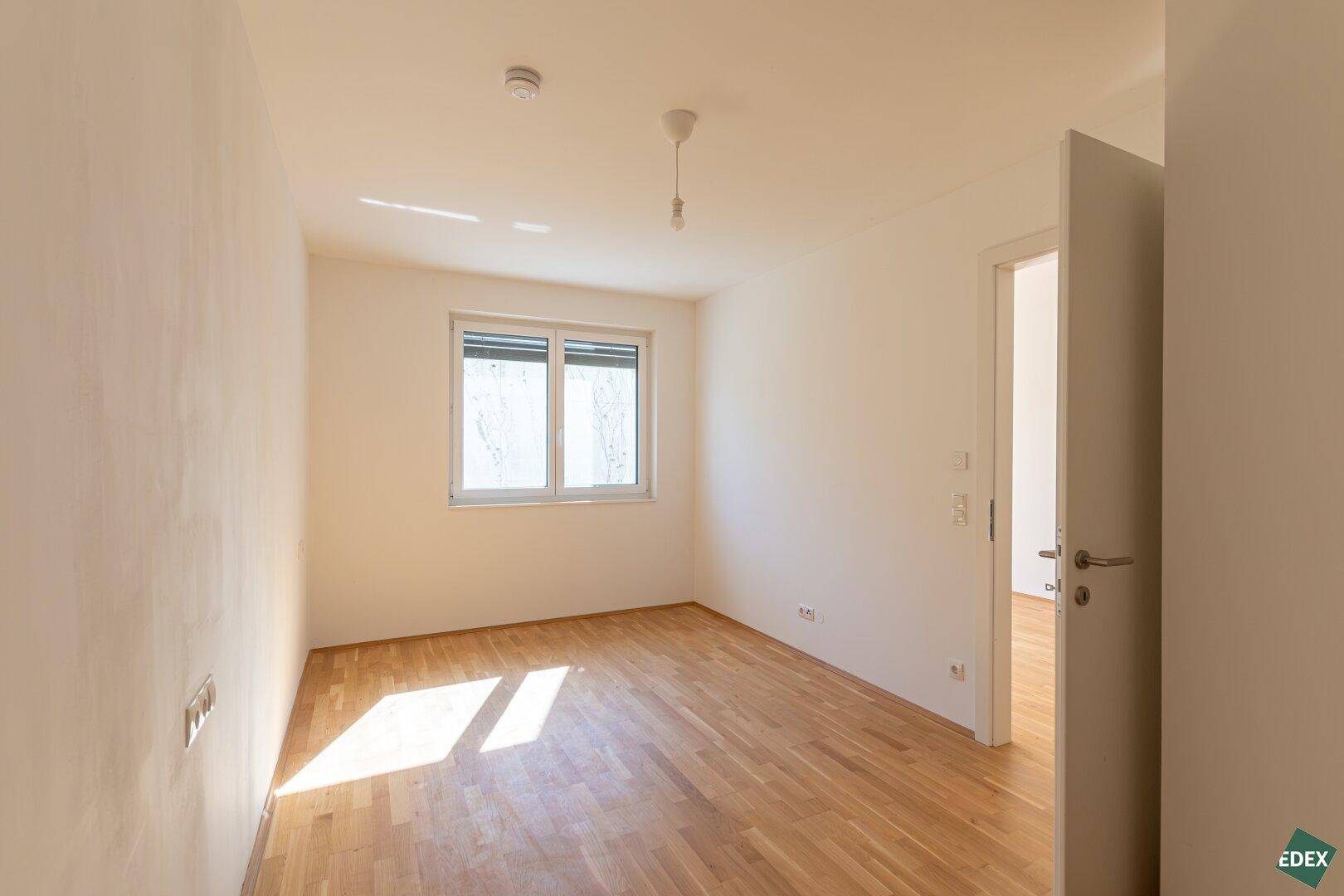 Helles, leeres Schlafzimmer mit Holzboden und Fenster, das viel Tageslicht hereinlässt.