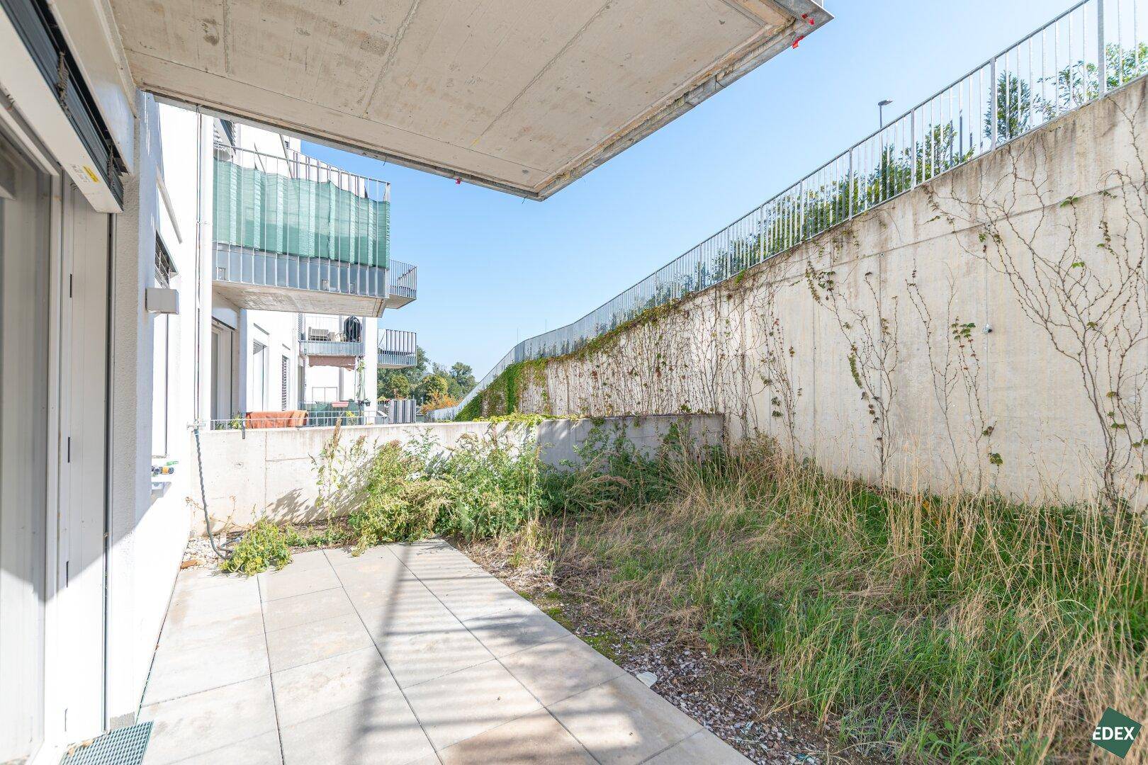 Kleine gepflasterte Terrasse mit angrenzender Grünfläche und Betonmauer im Außenbereich.