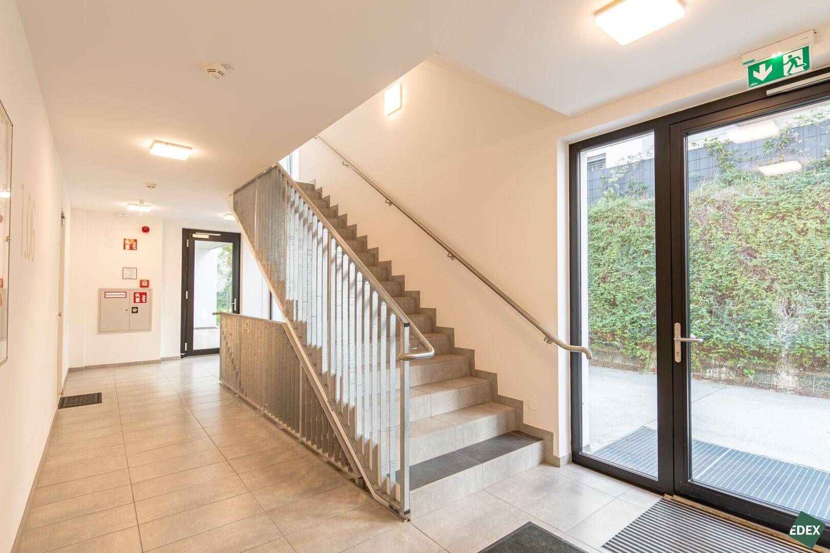 Heller Eingangsbereich mit Treppenhaus, Fliesenboden und großen Glastüren zum Außenbereich.
