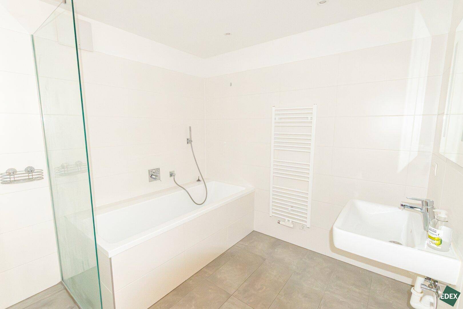 Modernes Badezimmer mit Badewanne, Dusche und beheiztem Handtuchhalter, in hellen Fliesen gehalten.