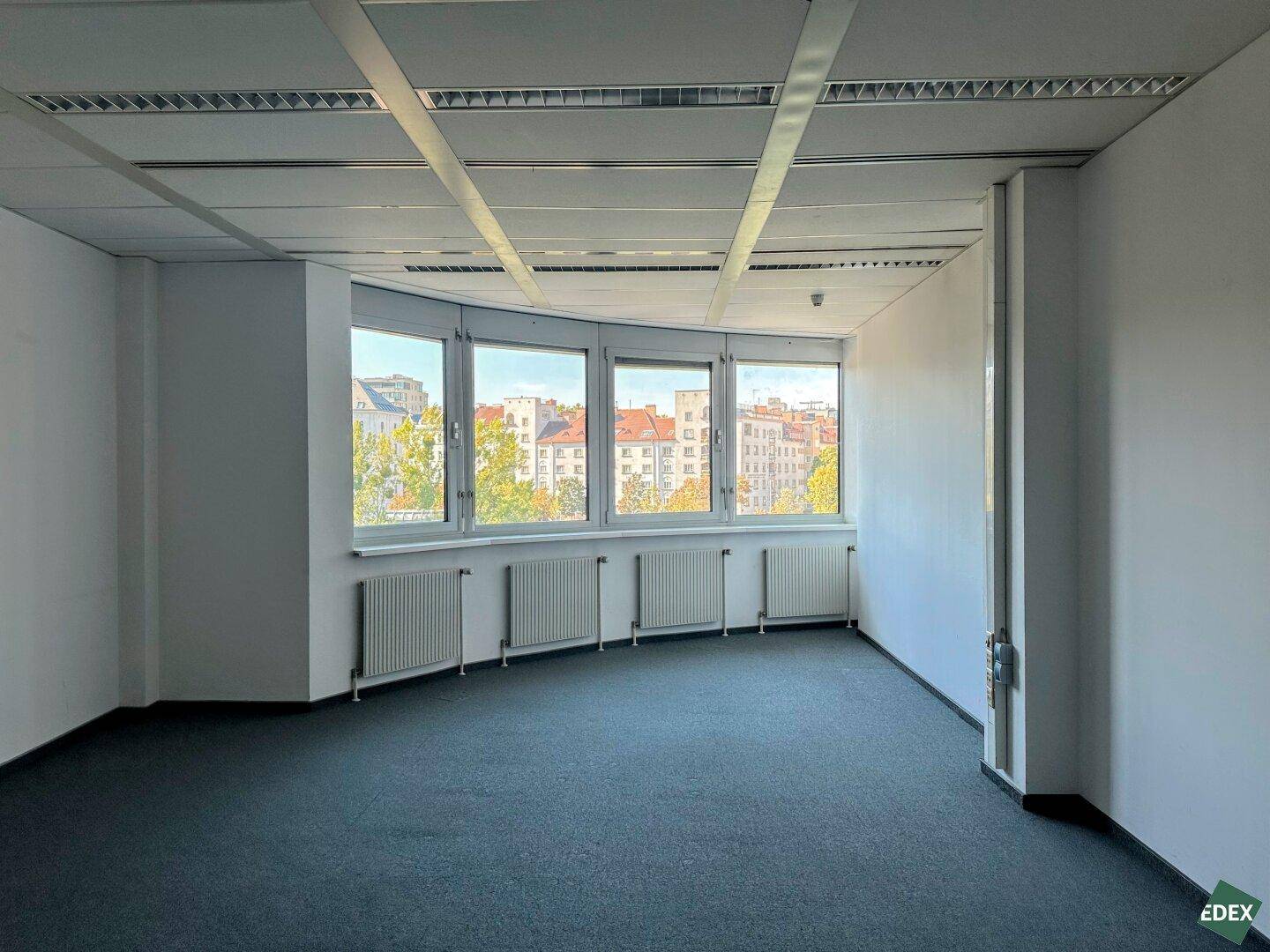 Heller Büroraum mit gebogener Fensterfront, Teppichboden und Heizkörpern unter den Fenstern.