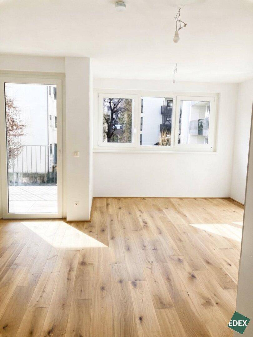 Leeres Wohnzimmer mit Holzboden und großen Fenstern, die viel Tageslicht hereinlassen.
