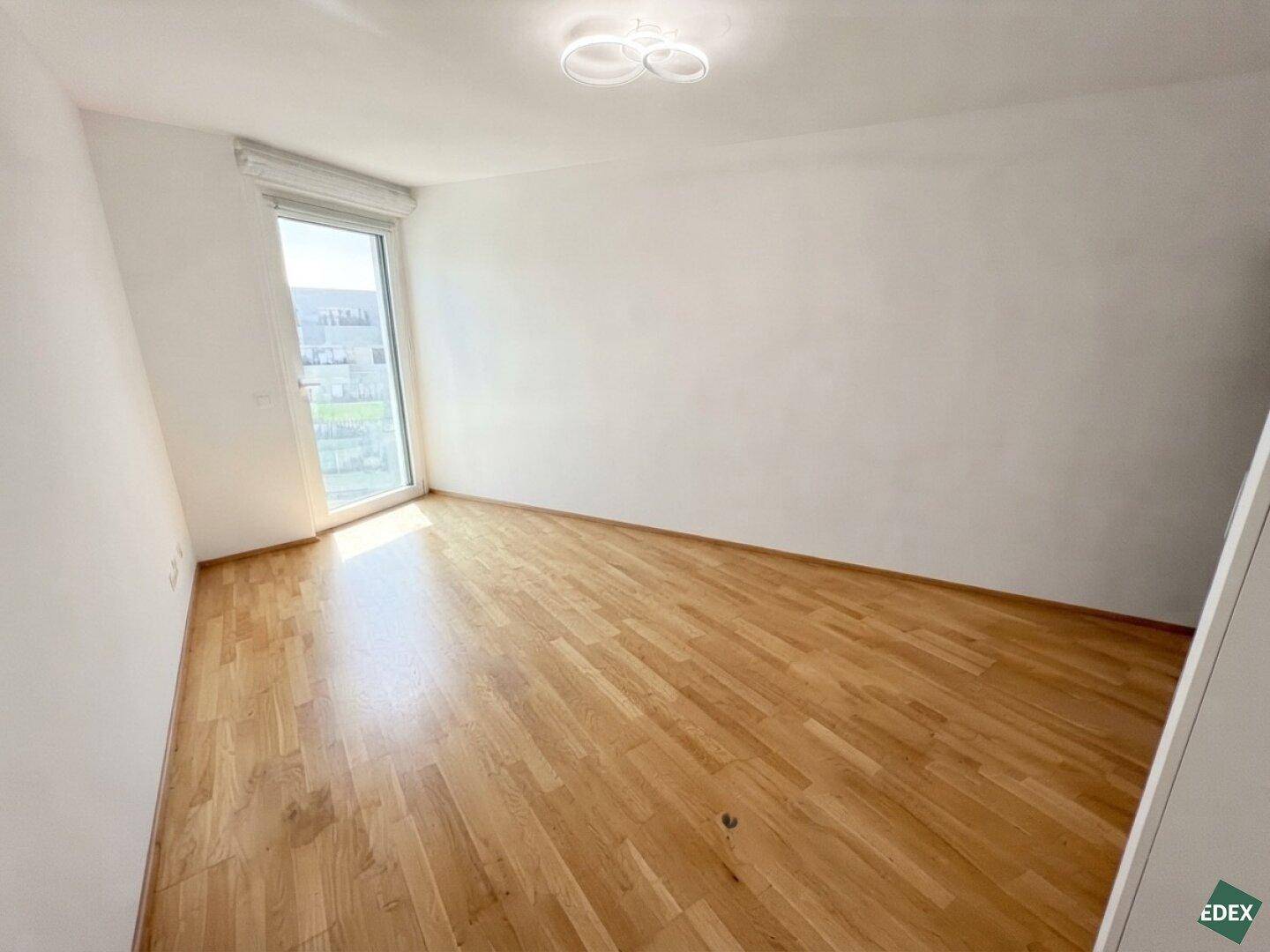 Helles Schlafzimmer mit Holzboden und großem Fenster, das viel Tageslicht hereinlässt.