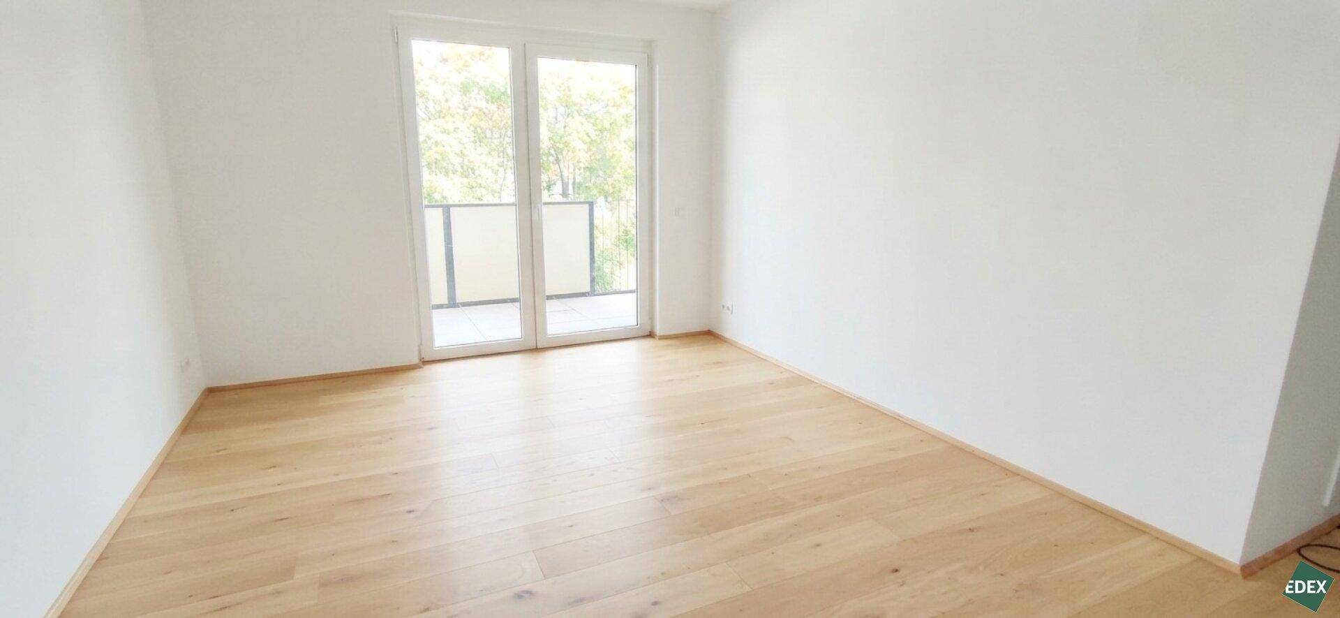 Leeres Wohnzimmer mit hellem Parkettboden und großen Fenstern zum Balkon.