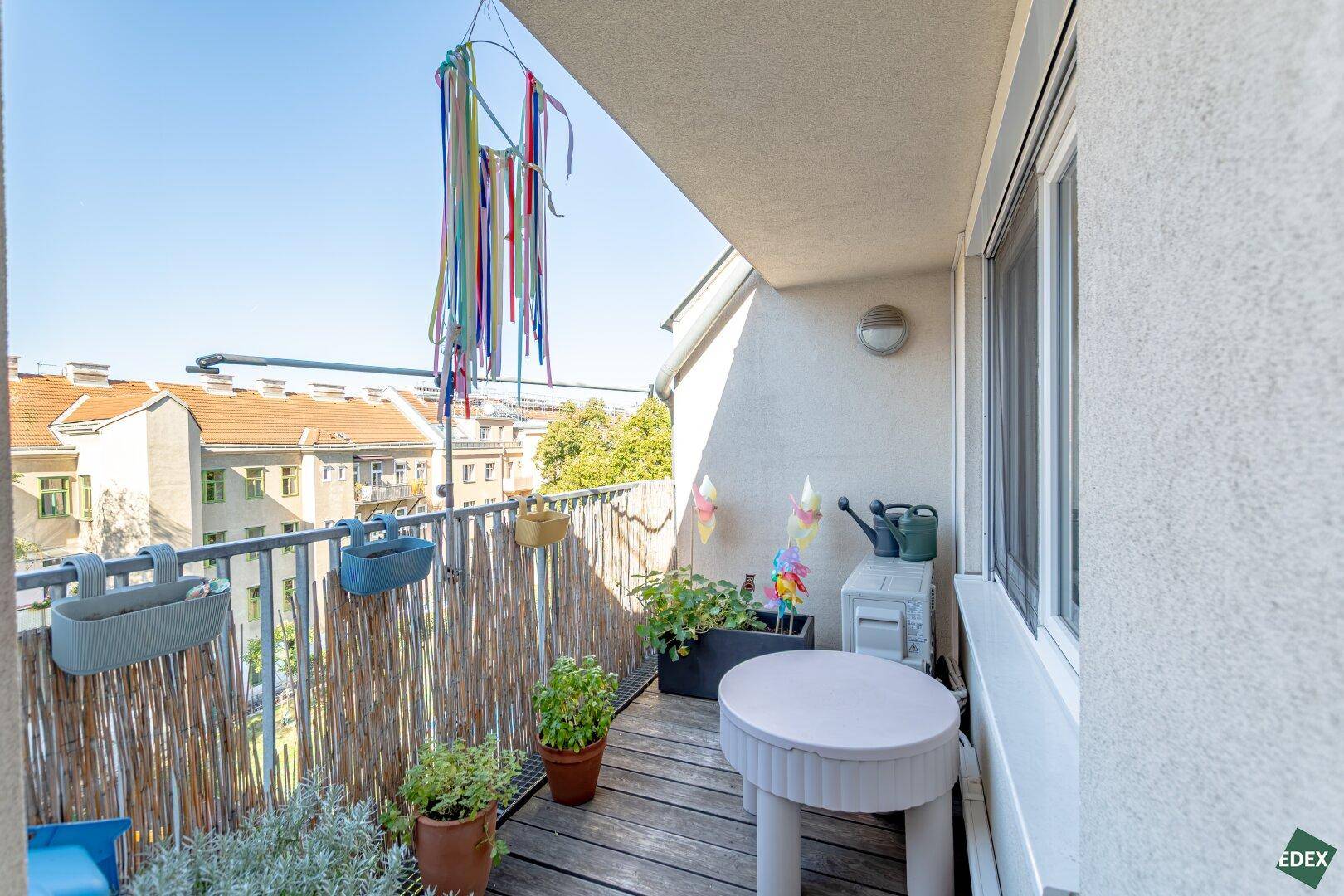 Kleiner, gemütlicher Balkon mit Holzboden, Pflanzen und Blick auf die umliegenden Gebäude.