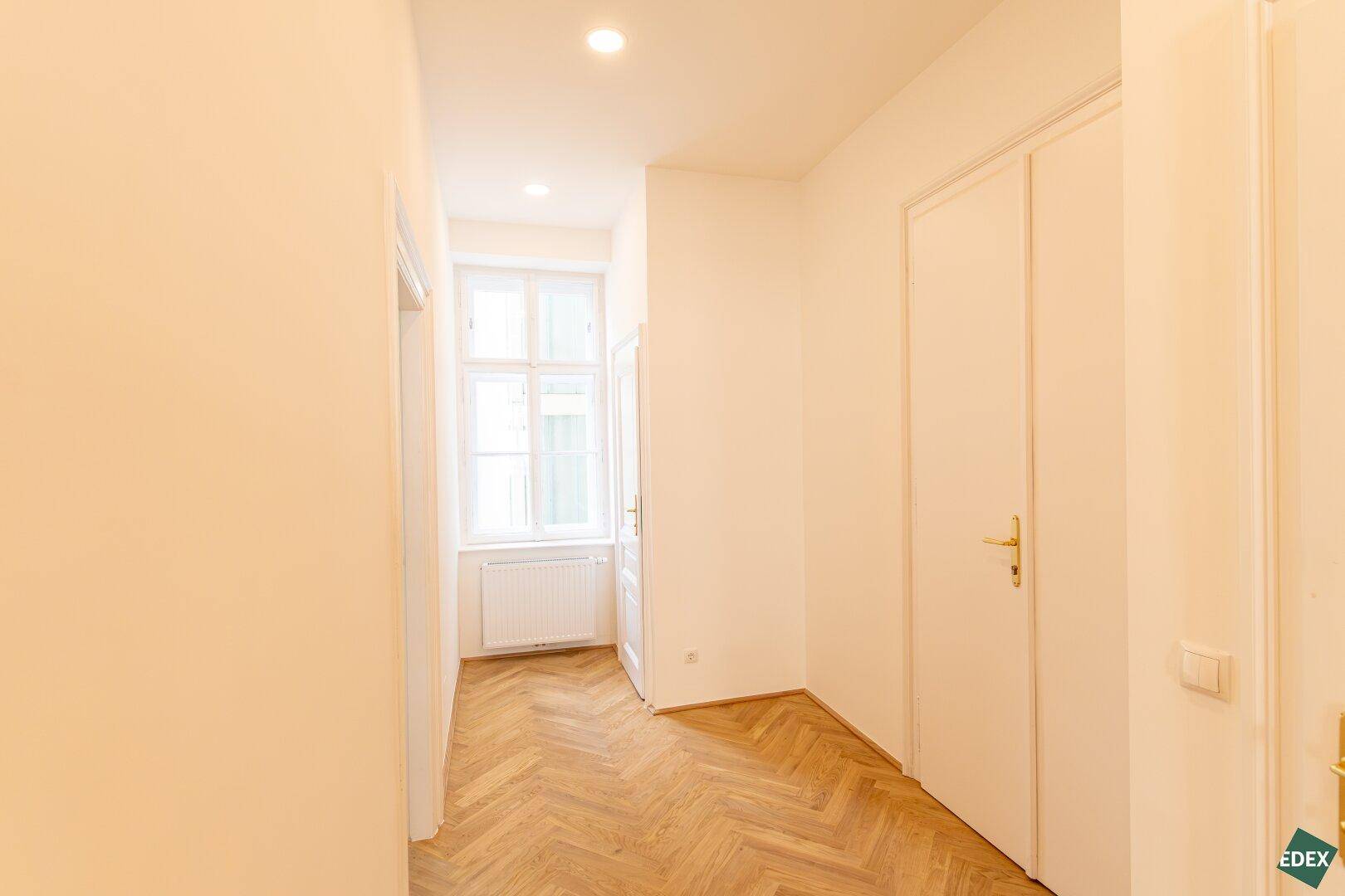 Langer Flur mit Fischgrätparkett und Fenster am Ende, sorgt für natürliche Belichtung.