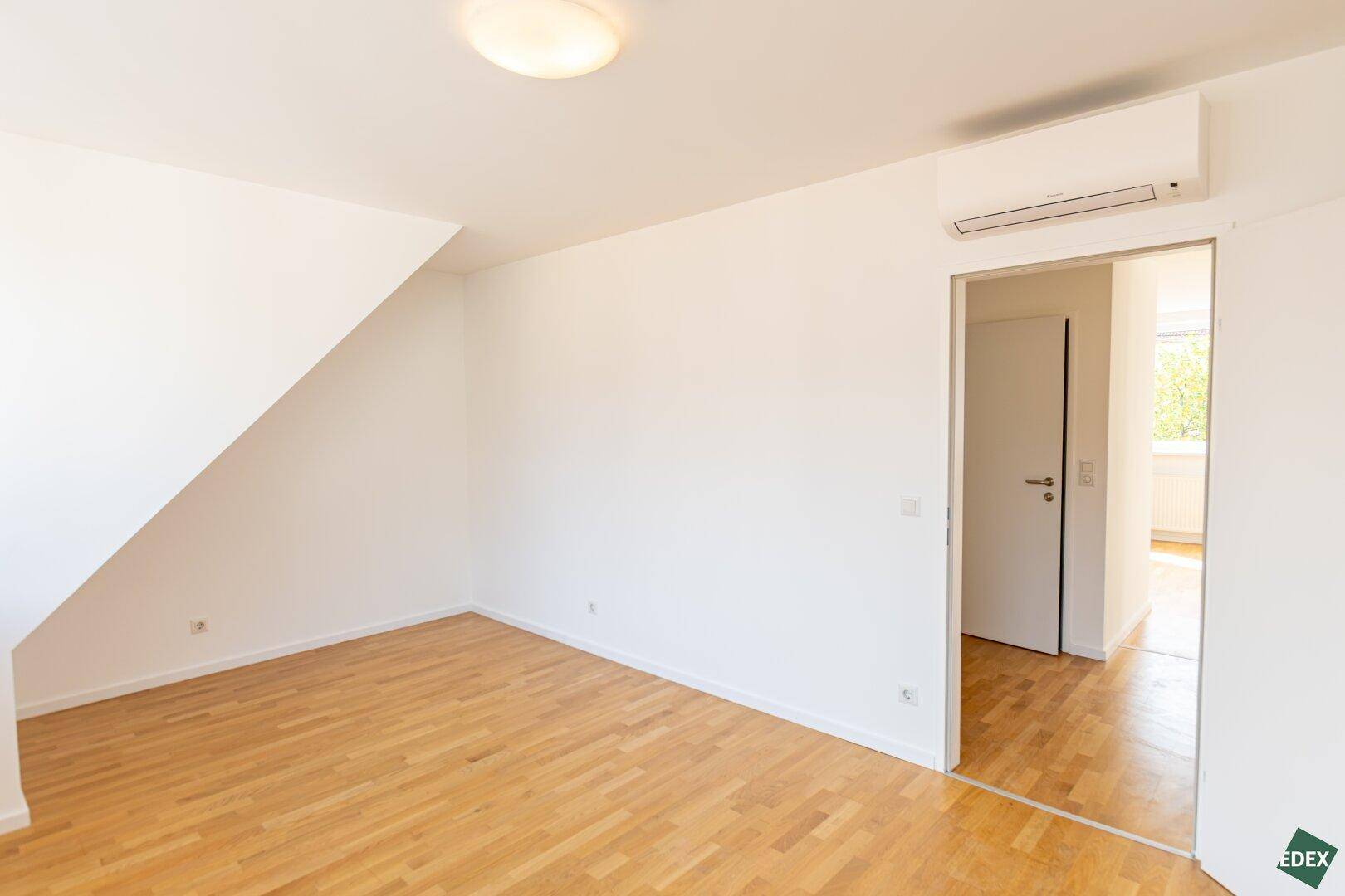 Heller Raum mit Holzparkettboden, Dachschräge und Zugang zu einem weiteren Zimmer.