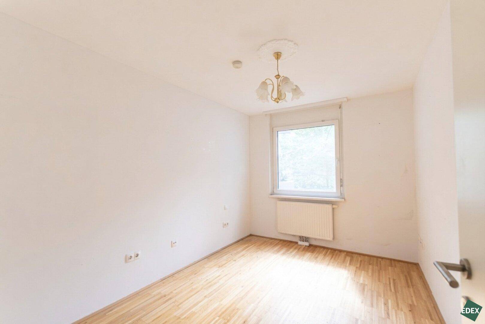 Helles, leeres Zimmer mit Parkettboden und einem Fenster, das Tageslicht hereinlässt.