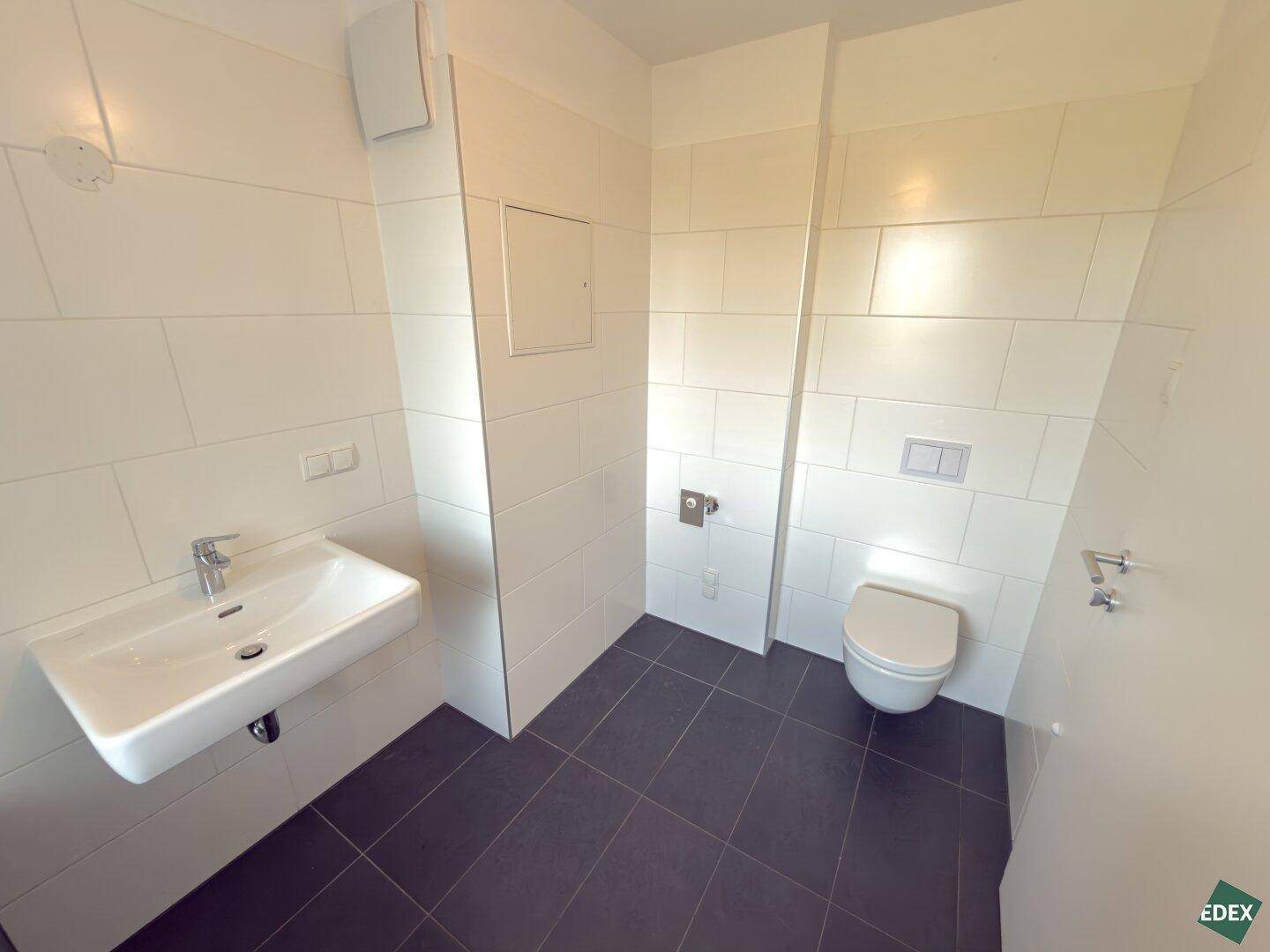 Modernes WC mit wandhängender Toilette, Einzelwaschbecken und Fliesen in Weiß und Dunkelgrau.