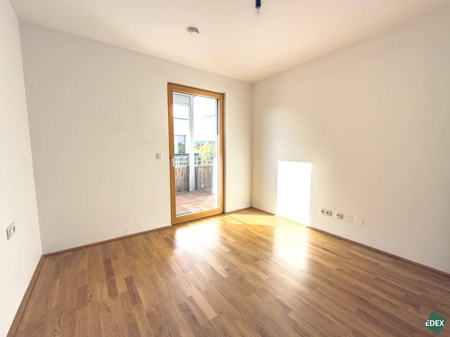 Helles Zimmer mit Holzboden und Zugang zu einem Balkon, durchflutet von natürlichem Sonnenlicht.