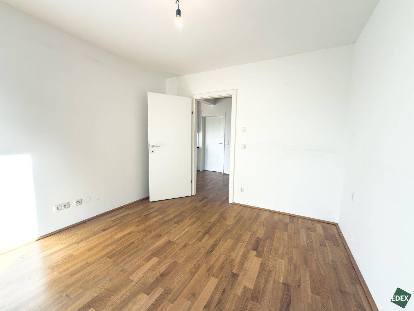Helles, leeres Zimmer mit Holzboden und weiß gestrichenen Wänden, mit Zugang zum Flur.