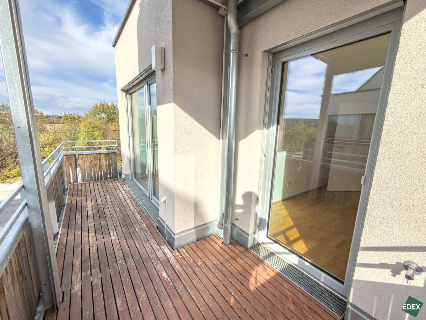 Geräumiger Balkon mit Holzbelag und Blick auf die grüne Umgebung und angrenzende Gebäude.