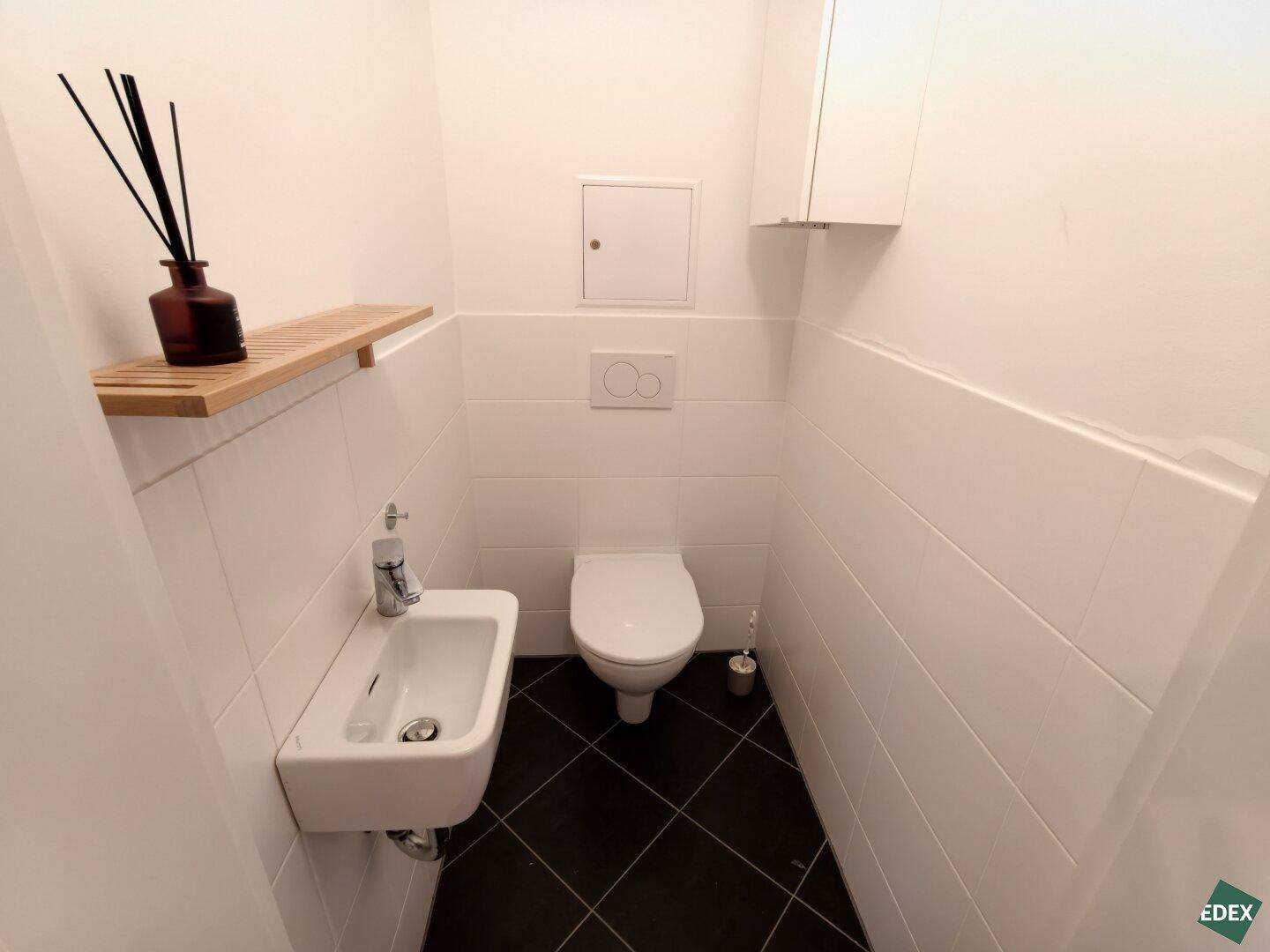 Separates Gäste-WC mit Wand-WC, kleinem Waschbecken und dunklen Bodenfliesen.