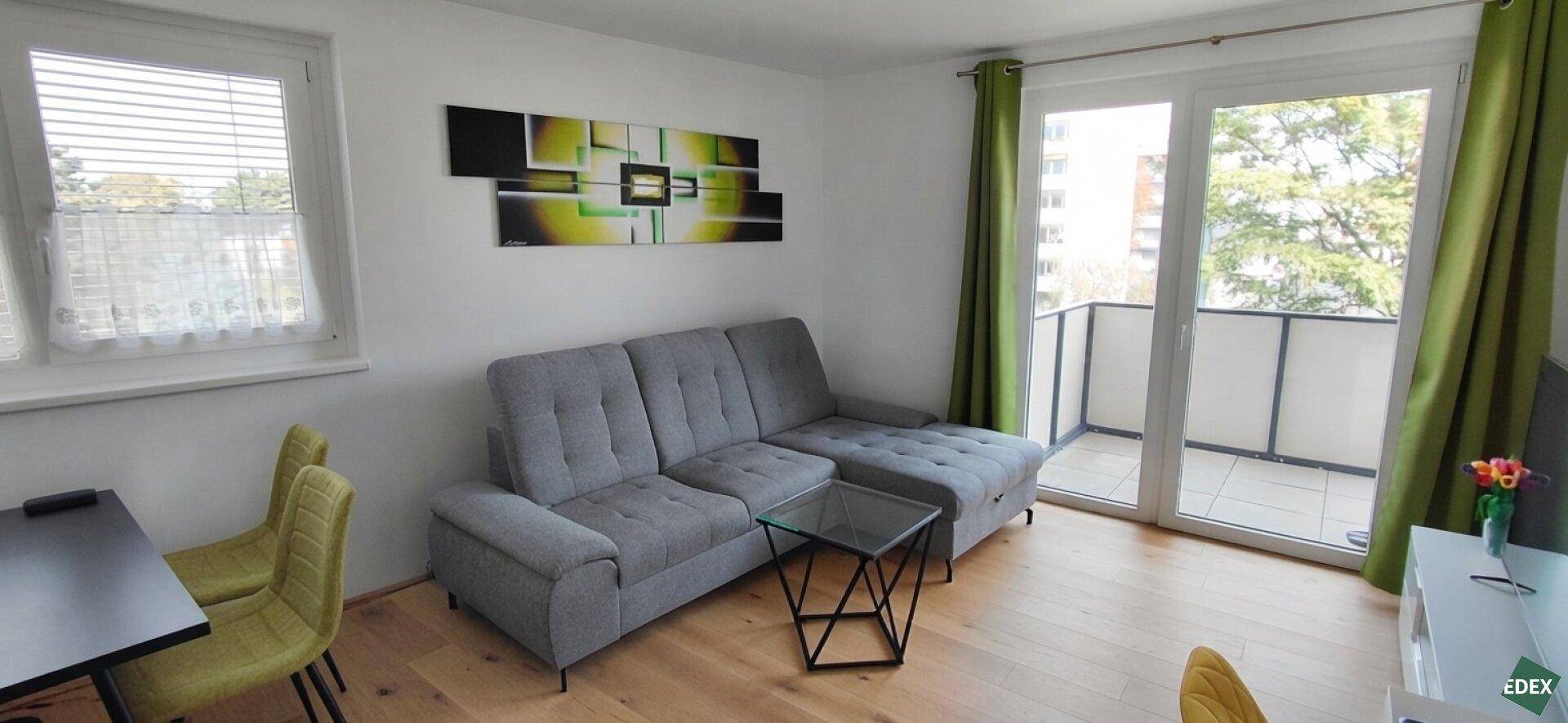 Gemütliches Wohnzimmer mit grauem Ecksofa, Holzboden und Balkonzugang.