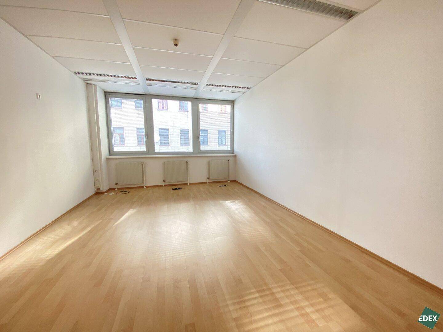 Helles, unmöbliertes Büro mit glänzendem Holzfußboden und großen Fenstern für natürliche Beleuchtung.