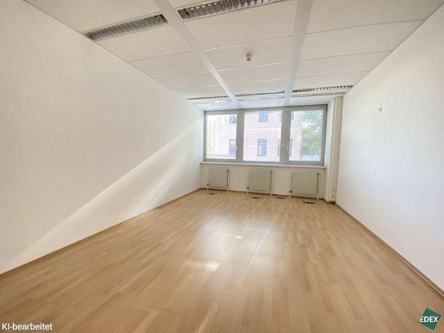 Leeres Büro mit hellem Holzfußboden, weißen Wänden und drei Fenstern, die viel Licht hereinlassen.