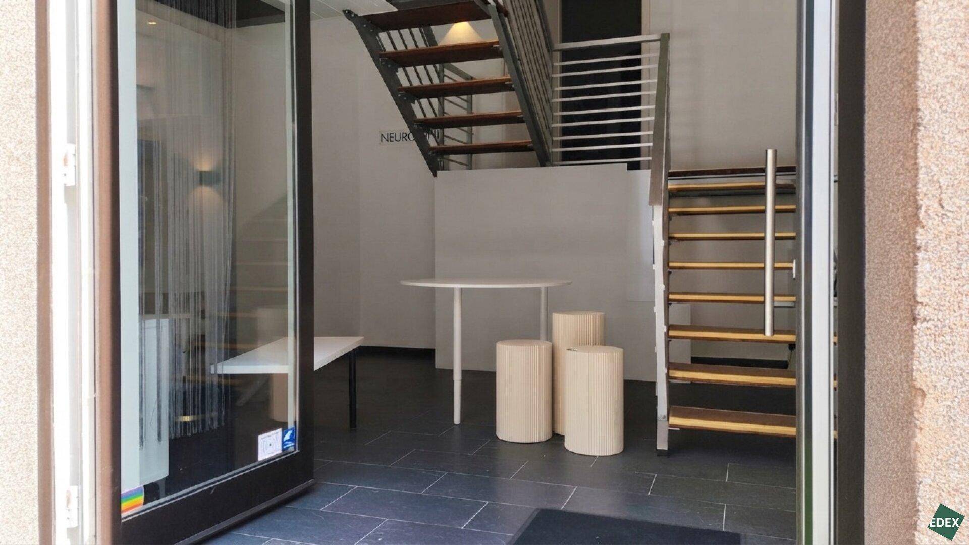 Eingangsbereich eines Gewerberaums mit offener Treppe, dunklem Fliesenboden und großen Glasfronten.
