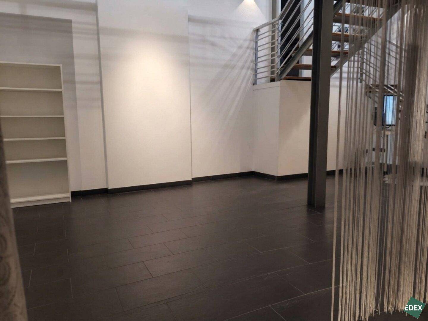 Großer, leerer Gewerberaum mit dunklem Fliesenboden, weiß gestrichenen Wänden und offener Treppe.