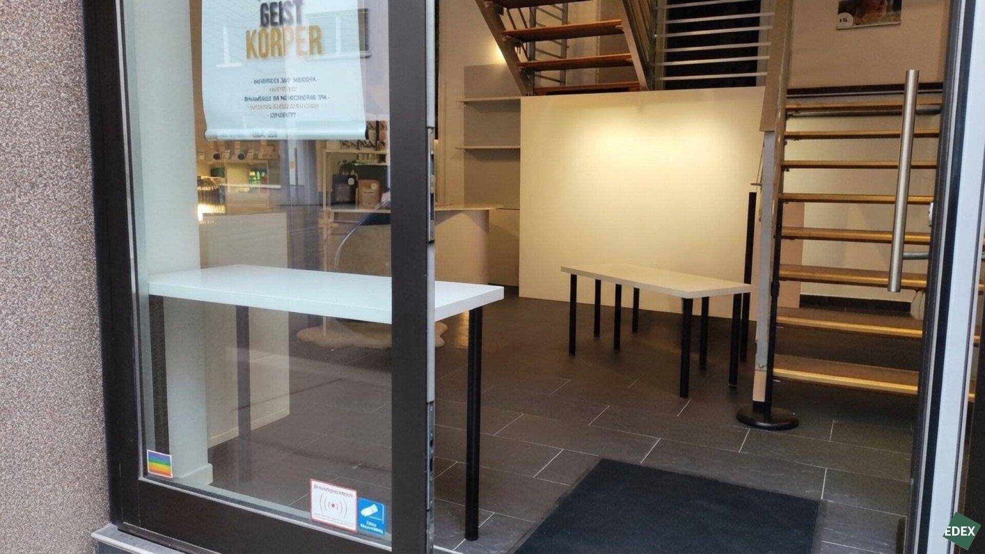 Eingangsbereich eines Geschäfts mit großen Glasfronten, dunklem Fliesenboden und offener Treppe.