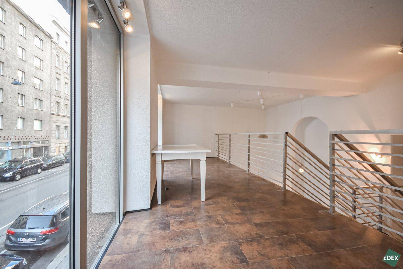 Obergeschoss eines Gewerberaums mit großen Fenstern, Blick auf die Straße und offener Treppe.