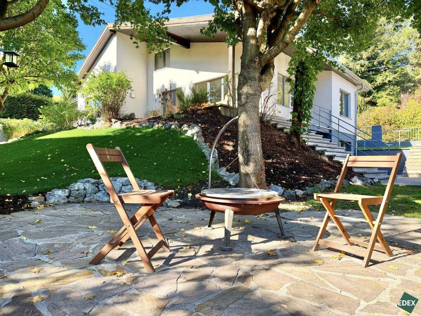 Gemütlicher Sitzbereich im Garten mit Feuerstelle und Stühlen unter schattenspendenden Bäumen.