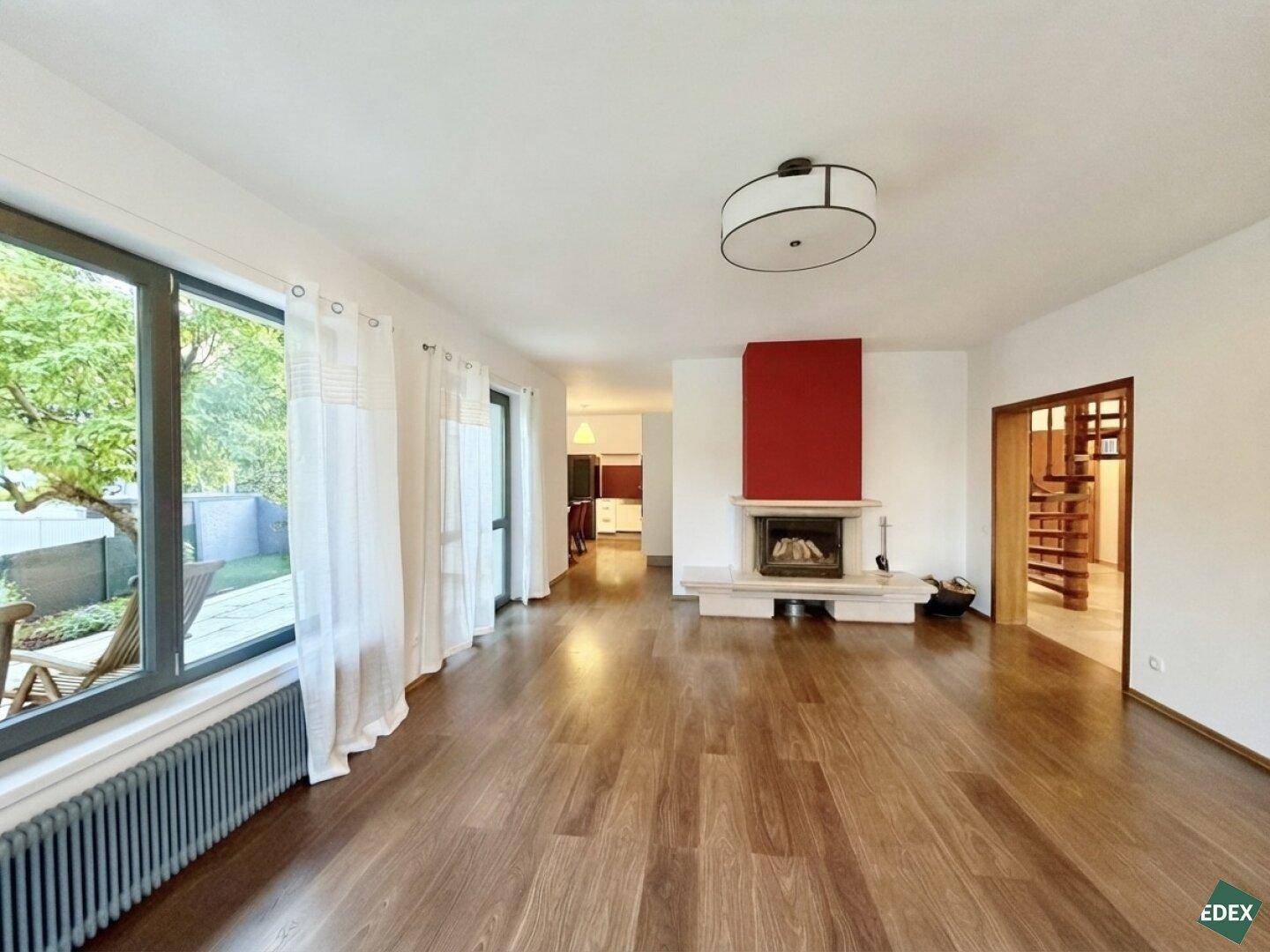 Geräumiges Wohnzimmer mit Holzboden, Kamin und großen Fenstern mit Blick ins Grüne.