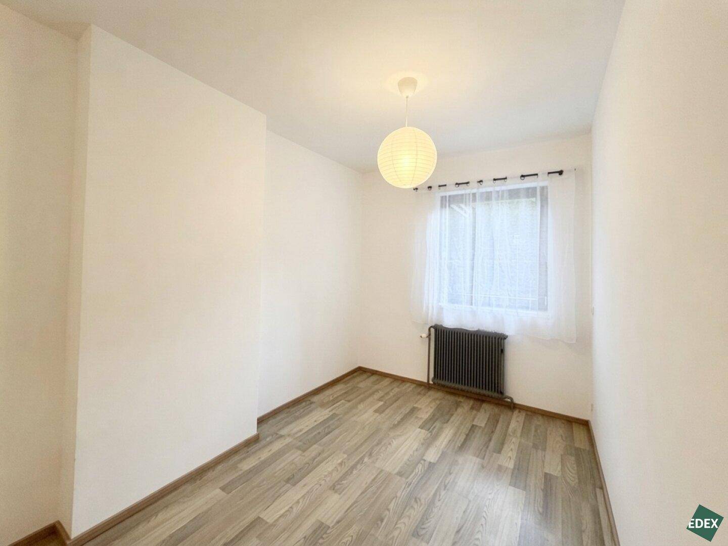 Leeres Schlafzimmer mit Laminatboden und einem Fenster, das viel Tageslicht hereinlässt.
