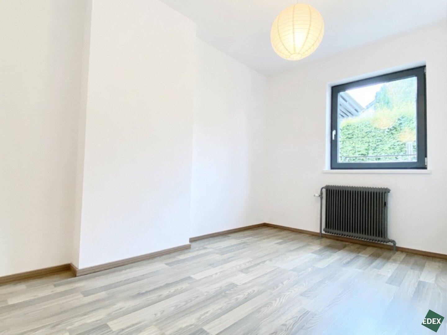 Schlafzimmer mit Laminatboden, weiß gestrichenen Wänden und Blick ins Grüne durch das Fenster.