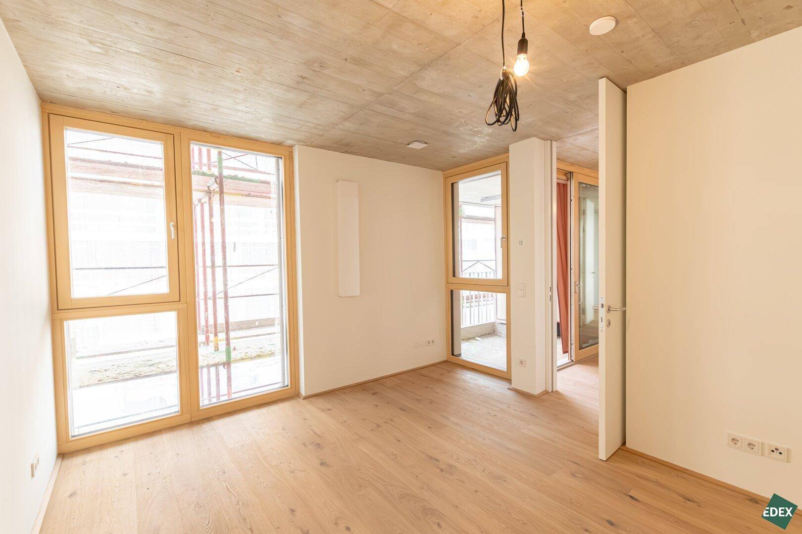 Heller, unmöblierter Raum mit Holzboden, Sichtbetondecke und großen Fenstern in einem Neubau.