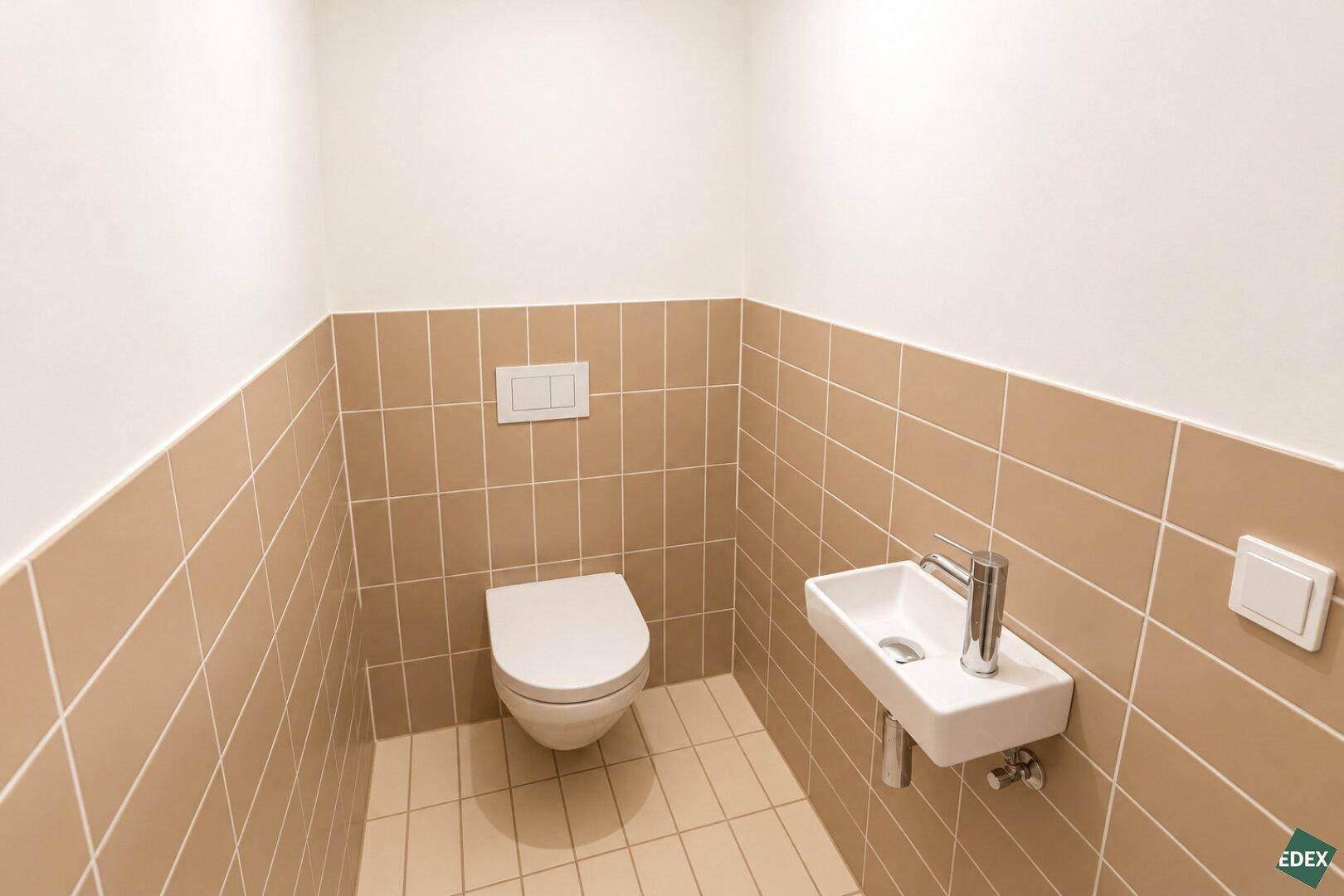Kleines Gäste-WC mit beigefarbenen Fliesen, wandhängendem WC und einem Handwaschbecken.