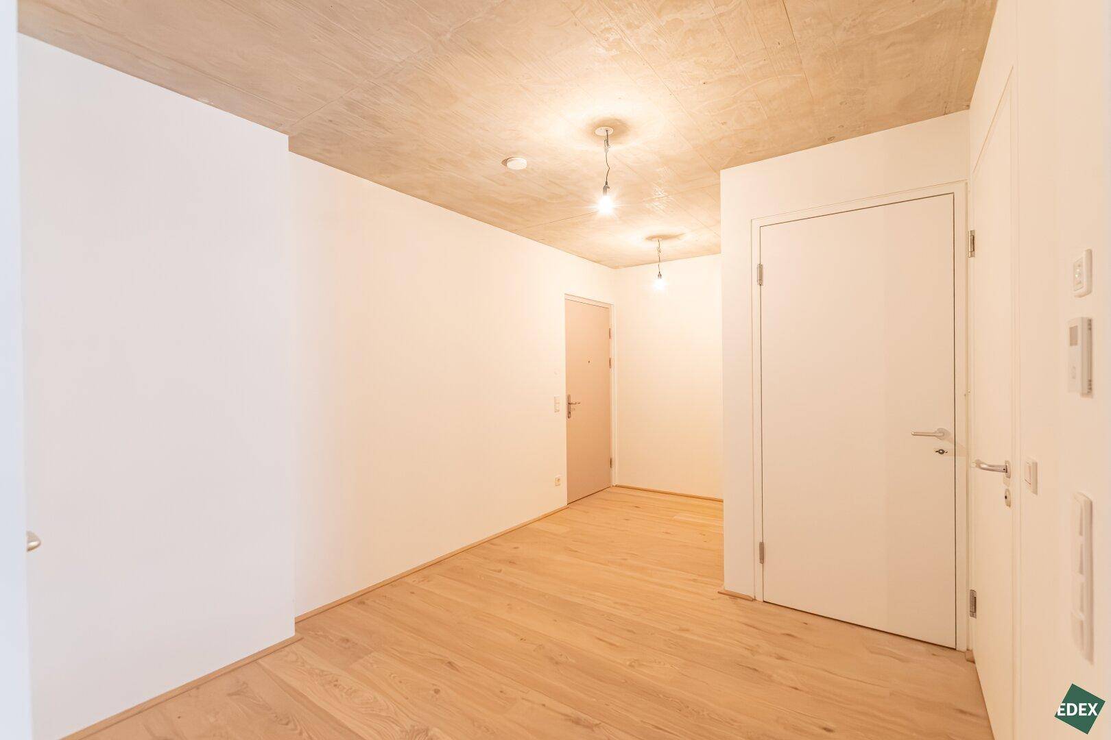 Langer, heller Flur mit Holzboden, Sichtbetondecke und mehreren Türen zu den Zimmern.