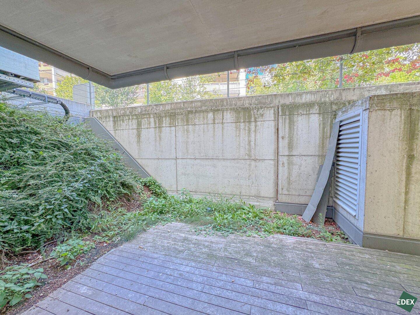 Eine schattige Holzterrasse im Freien, umgeben von Betonwänden und üppiger Vegetation.