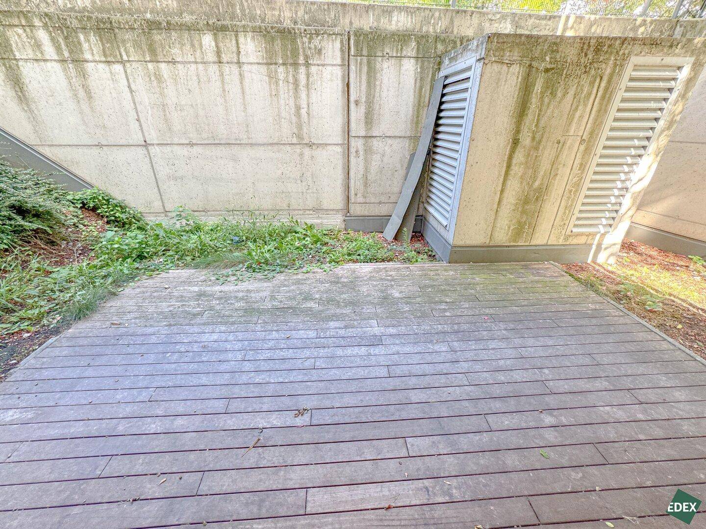 Eine breite Holzterrasse im Freien, umgeben von Betonmauern und etwas Grün, bietet Platz zum Entspannen.